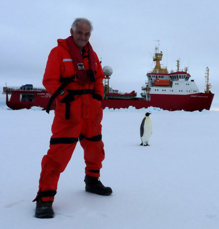 Photo of David Meldrum in Antarctica with two penguins and the research vessel in the background