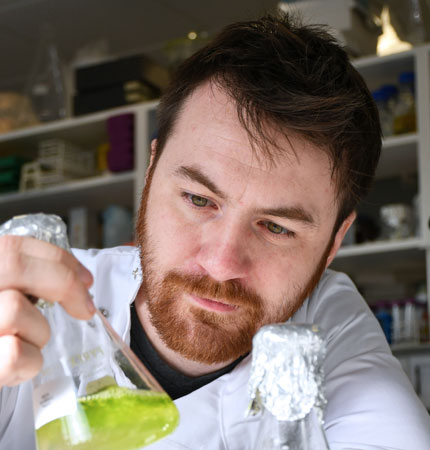 Head and shoulder photo of Mikey Ross in a white lab coat with a flask full of algae