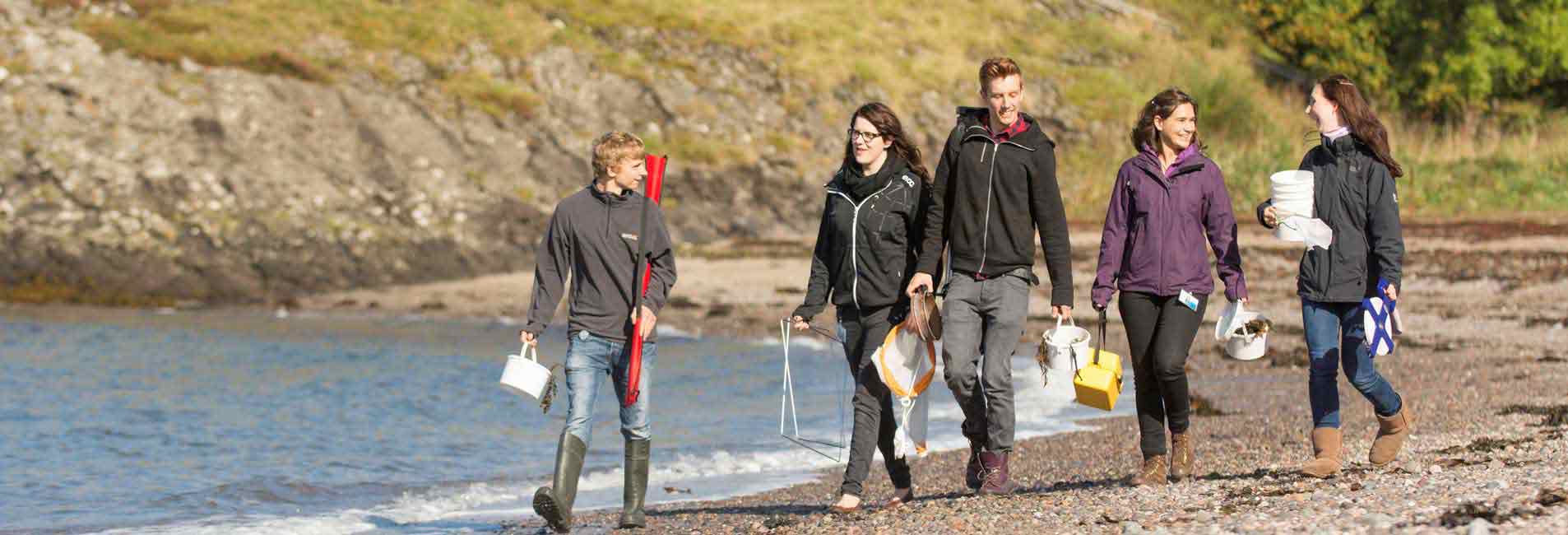 The picture shows a group of fresh-faced marine science undergraduate students walking on the beach with some of their fieldwork equipment