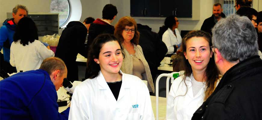 Photo showing a parent talking to two current students in lab coats while in the background others look through microscopes etc