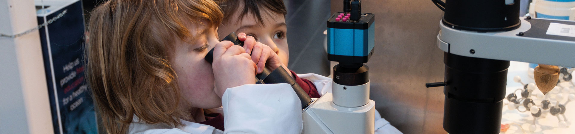 Two young learners in lab coats look through a microscope 