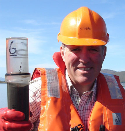 
        Photo of Professor Black in orange hard hat and swim vest with sediment core during sampling trip
        