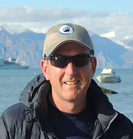 Gillen Wood wearing a cap and sunglasses with boats and mountains behind him