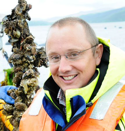 
        Adam Hughes on a marine farm with rope-grown mussels in the background
        