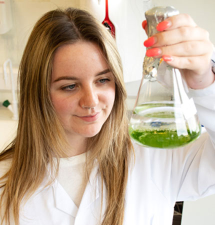 Female support scientist in lab coat holding a glass vial with green algae