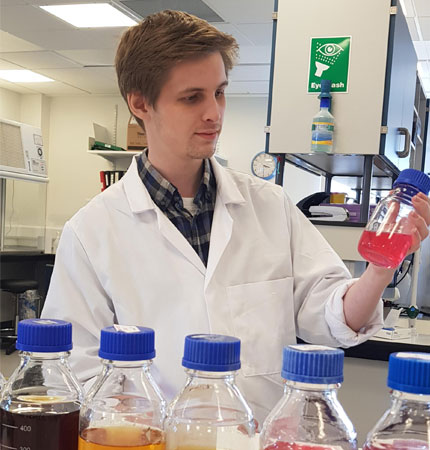 
        Jamie Rowell wearing a white lab coat in with an algal culture inside the Culture Collection lab
        