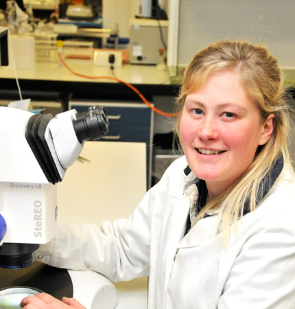 Naomi Thomas in white lab coat sitting on a microscope to identify toxic algae