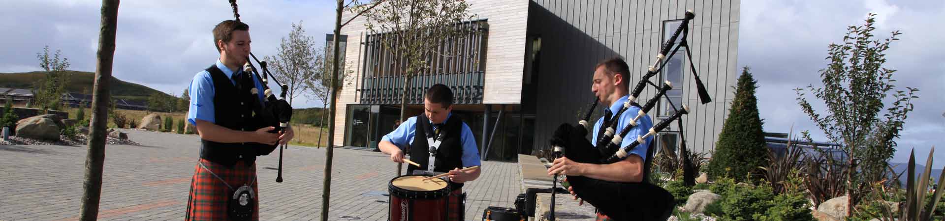 Image showing three pipers outside our campus preparing to pipe graduating students into the graduation marquee